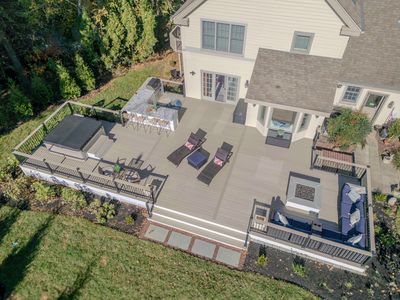 Aerial view of the Chadds Ford back deck showing the entire multi-zone outdoor living build: outdoor kitchen, recessed hot tub, wicker chaises, and curved blue sectional surrounding a square fire pit — Nature's Call hardscaping and outdoor living project