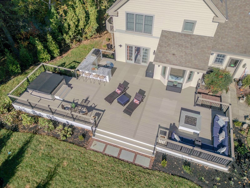 Aerial view of the Chadds Ford back deck showing the entire multi-zone outdoor living build: outdoor kitchen, recessed hot tub, wicker chaises, and curved blue sectional surrounding a square fire pit — Nature's Call hardscaping and outdoor living project
