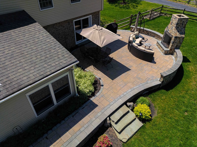 Aerial view of a Garnet Valley PA back patio: a curved paver platform surrounds an in-ground hot tub and full-height fieldstone outdoor fireplace, with a tan umbrella shading the dining area — Nature's Call landscaping and hardscaping project