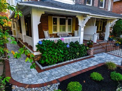 Front-yard view of a Wilmington Delaware row home with curved blue-gray paver walkway, red brick edging, brick-faced raised planters, and a hedge of blue and purple hydrangeas — Nature's Call hardscaping and front-entry rebuild